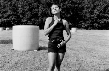 Woman in a black top, shorts and boots standing in a field with large round bales of hay, surrounded by trees, photographed in black and white.
